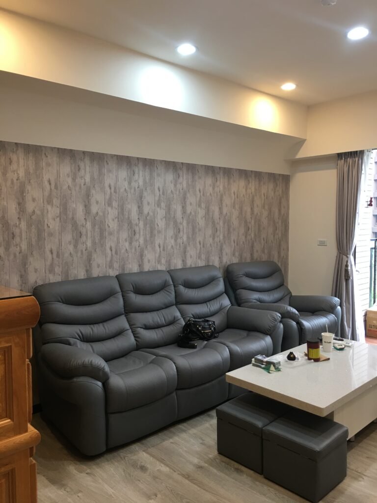 客廳沙發區室內設計，深灰色皮質沙發搭配仿木紋質感壁紙背景牆，展現現代沉穩風格。