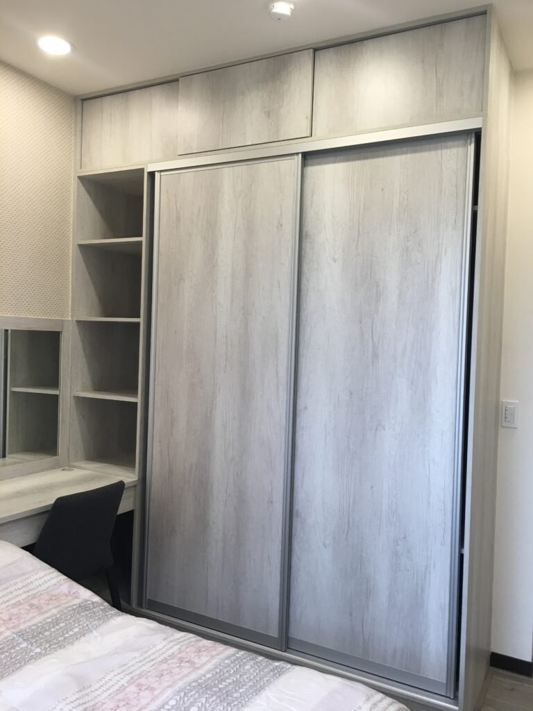 臥室系統家具設計，包含淺灰色木紋頂天立地推拉門衣櫃、開放式收納格，以及緊鄰床邊的一體化書桌兼化妝台。